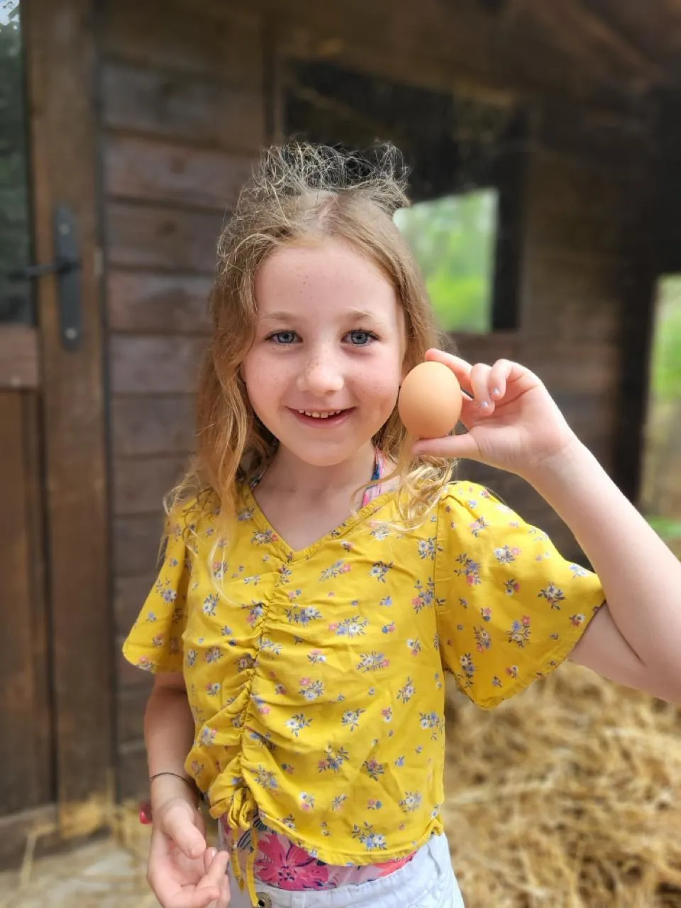 Child holding an egg, smiling outside wooden cabin.